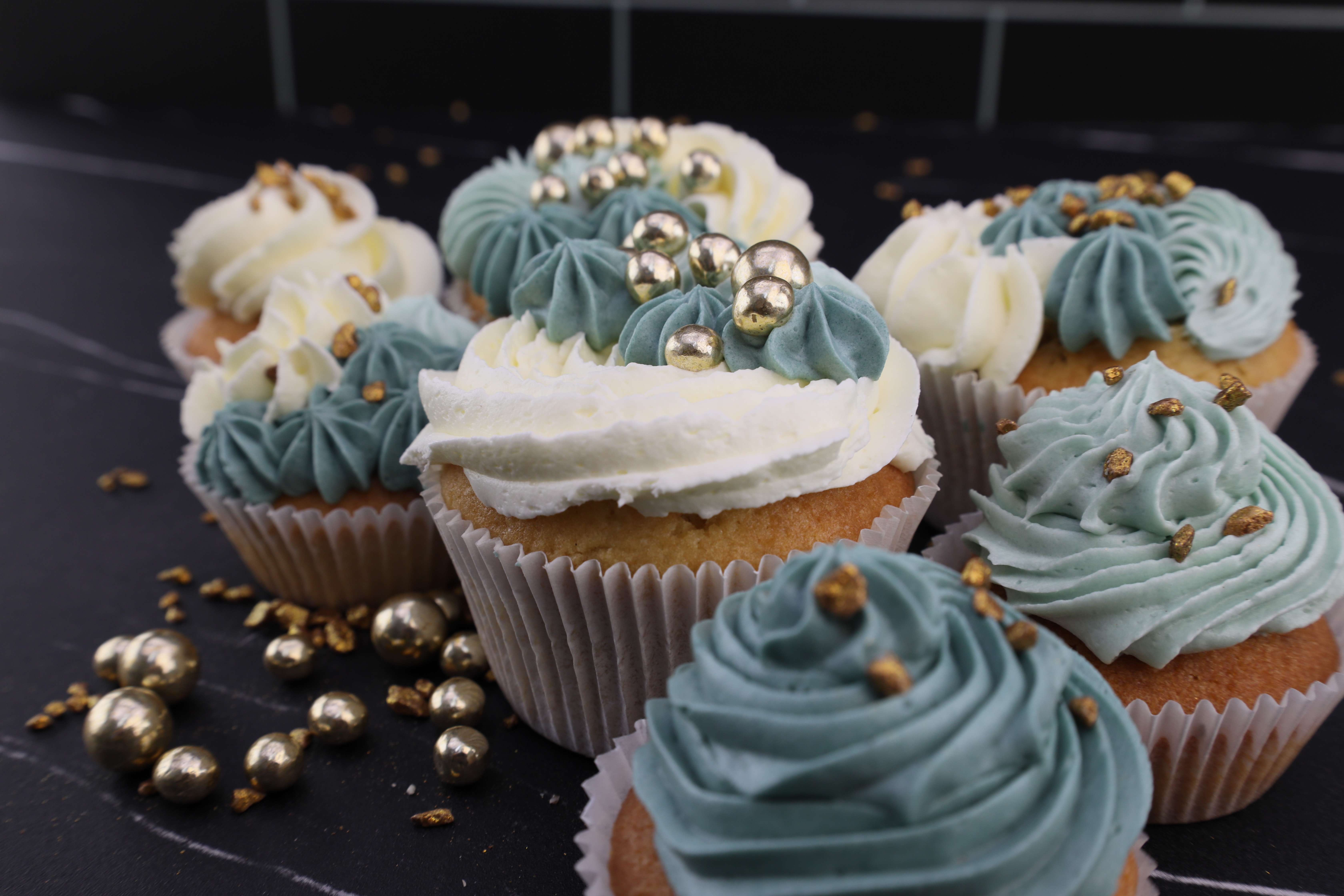 Köstlicher Muffins aus eukalyptusblauer und weißer Buttercreme mit goldenen essbaren Perlen und Goldzucker oben drauf. 